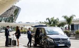 A black Mercedes-Benz V-Class minivan is parked at an airport, with three people standing beside it. Two are dressed in suits, one male and one female, each with a rolling suitcase. The third person, a man in a suit, is holding the sliding door open. The airport terminal, featuring a modern glass facade, is visible in the background along with palm trees and other greenery. The scene suggests a luxury airport transfer service.